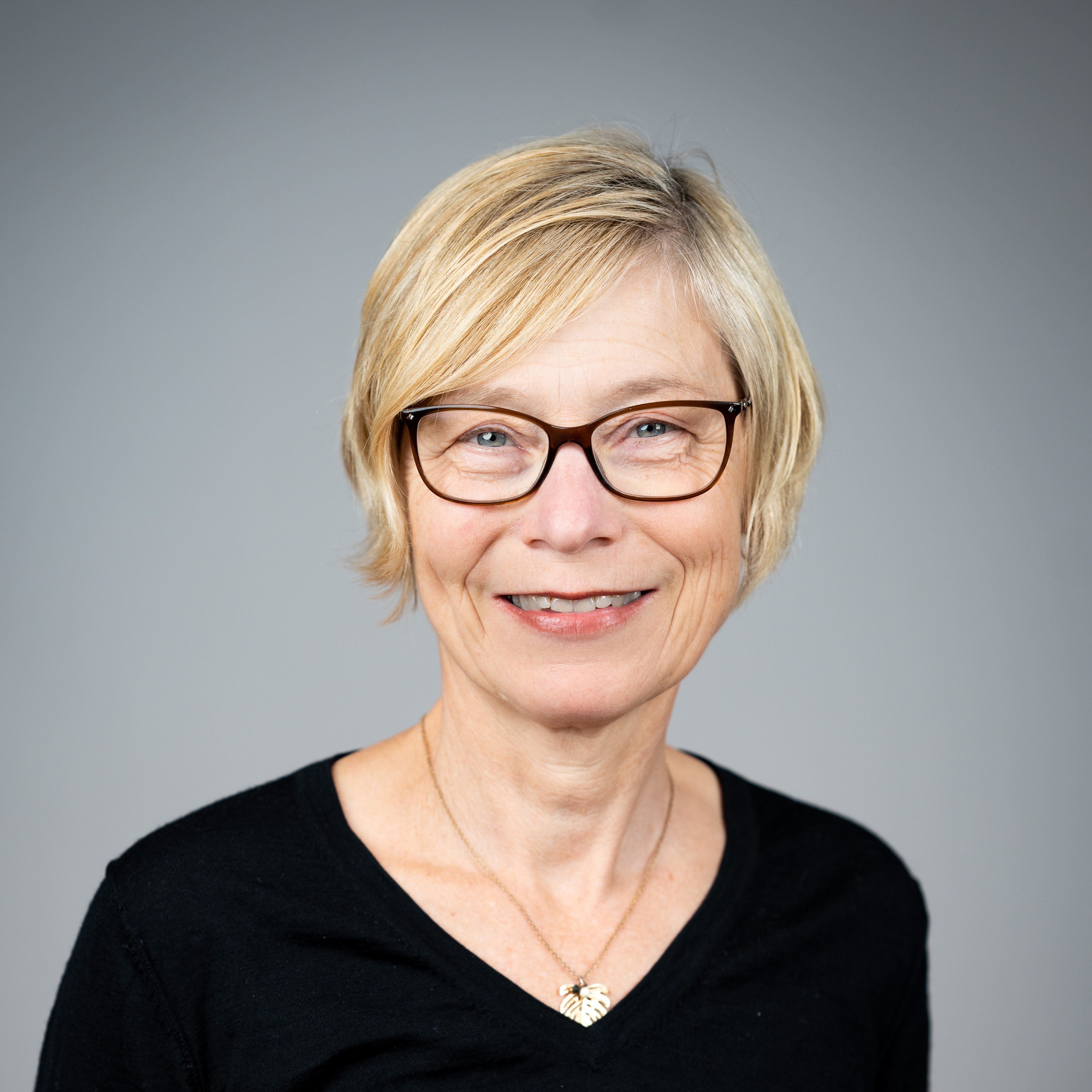 Jannecke Wiers-Jenssen smiler mot kamera i et portrettbilde, med kort blondt hår, briller og mørk topp mot en nøytral grå bakgrunn.