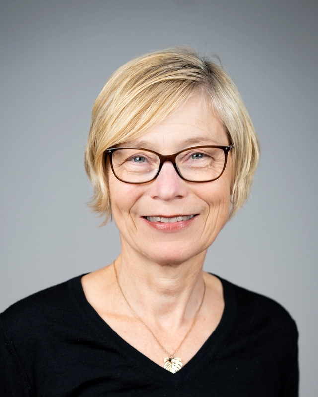 Jannecke Wiers-Jenssen smiler mot kamera i et portrettbilde, med kort blondt hår, briller og mørk topp mot en nøytral grå bakgrunn.