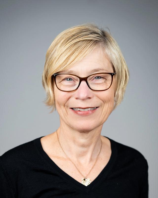 Jannecke Wiers-Jenssen smiler mot kamera i et portrettbilde, med kort blondt hår, briller og mørk topp mot en nøytral grå bakgrunn.
