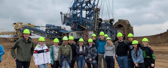 15 personer, kvinner og menn, står med anleggshjelmer foran en stor maskin.