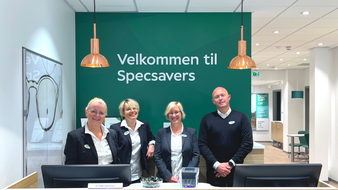 Tre kvinner og en mann står ved disken i en Specsavers-butikk med plakaten "Velkommen til Specsavers" bak seg.