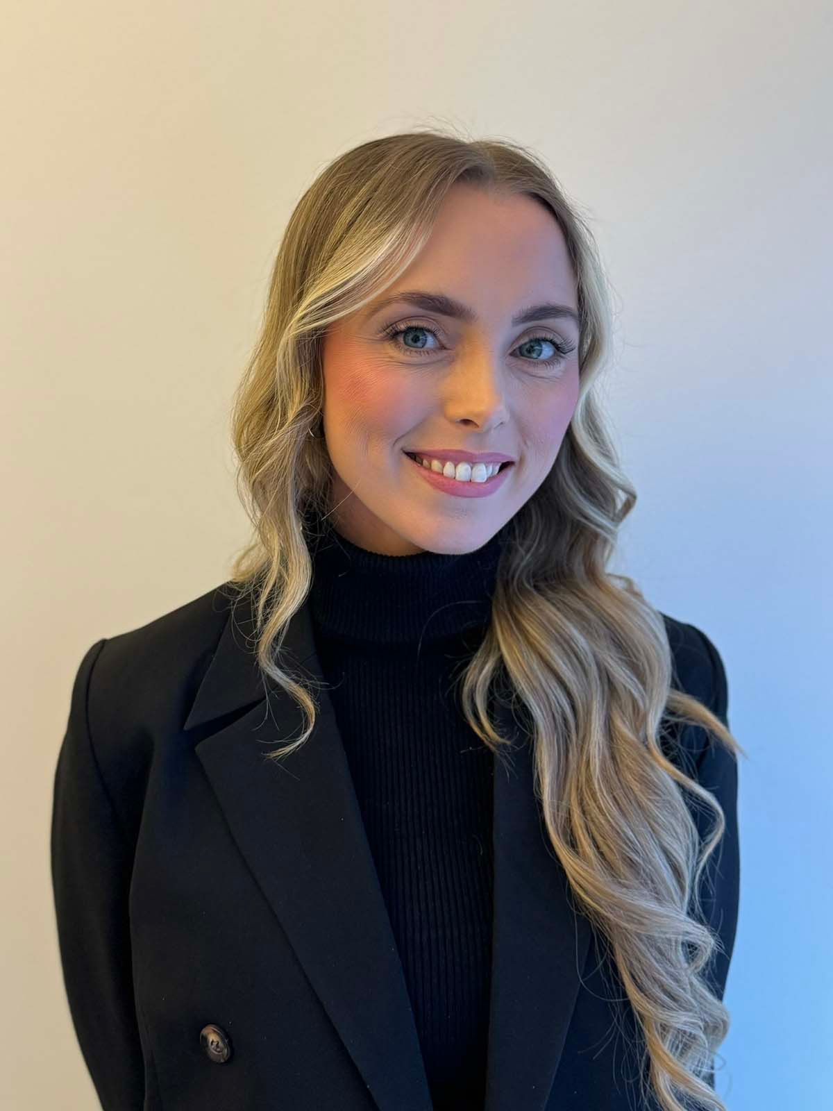 Portrett av en kvinne med langt blondt hår, iført svart blazer og sort høyhalset genser, mot en lys bakgrunn.