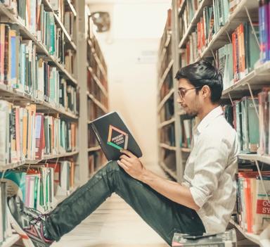 Student som sitter på gulvet mellom hylleradene på et bibliotek og leser en bok. 