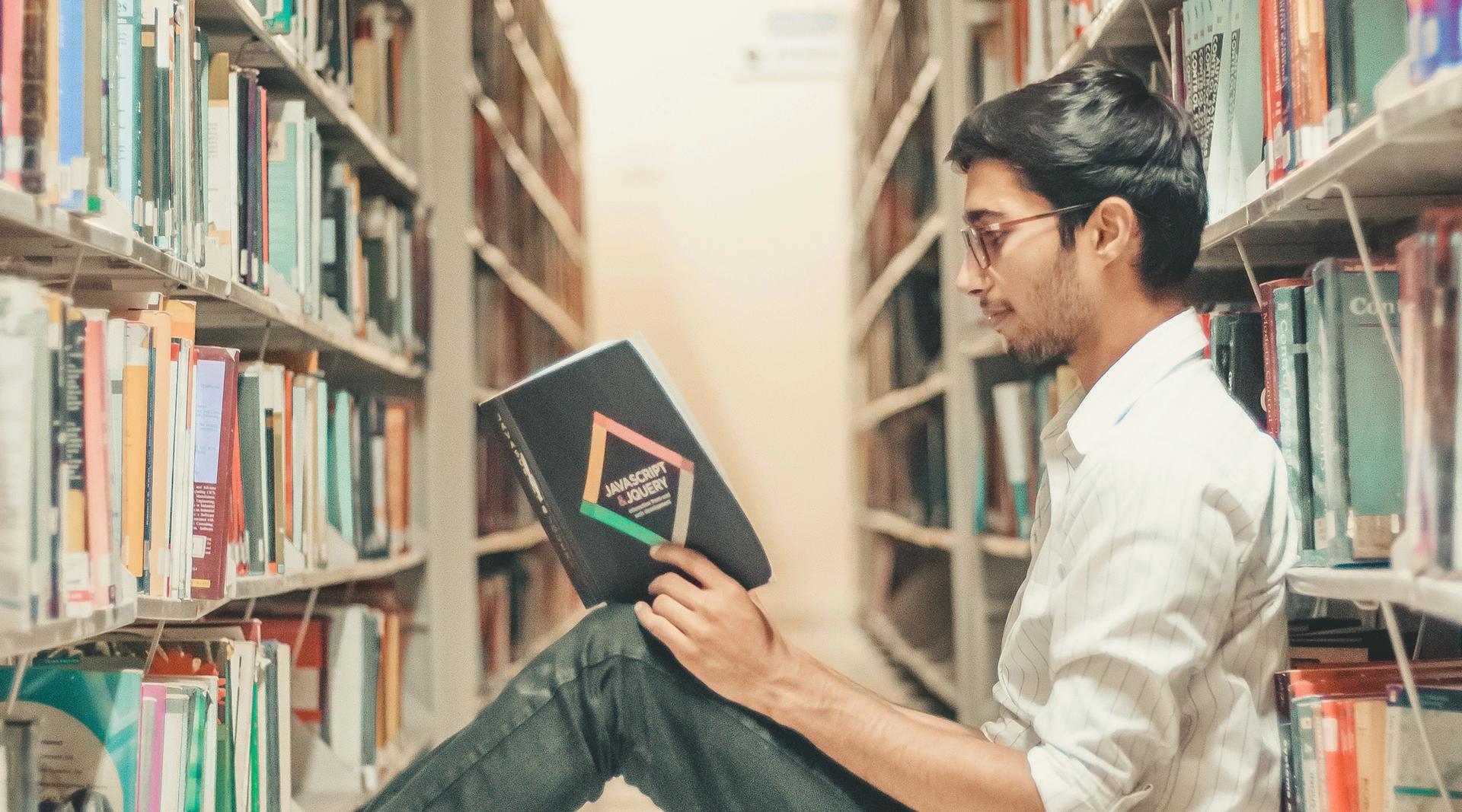 Student som sitter på gulvet mellom hylleradene på et bibliotek og leser en bok.