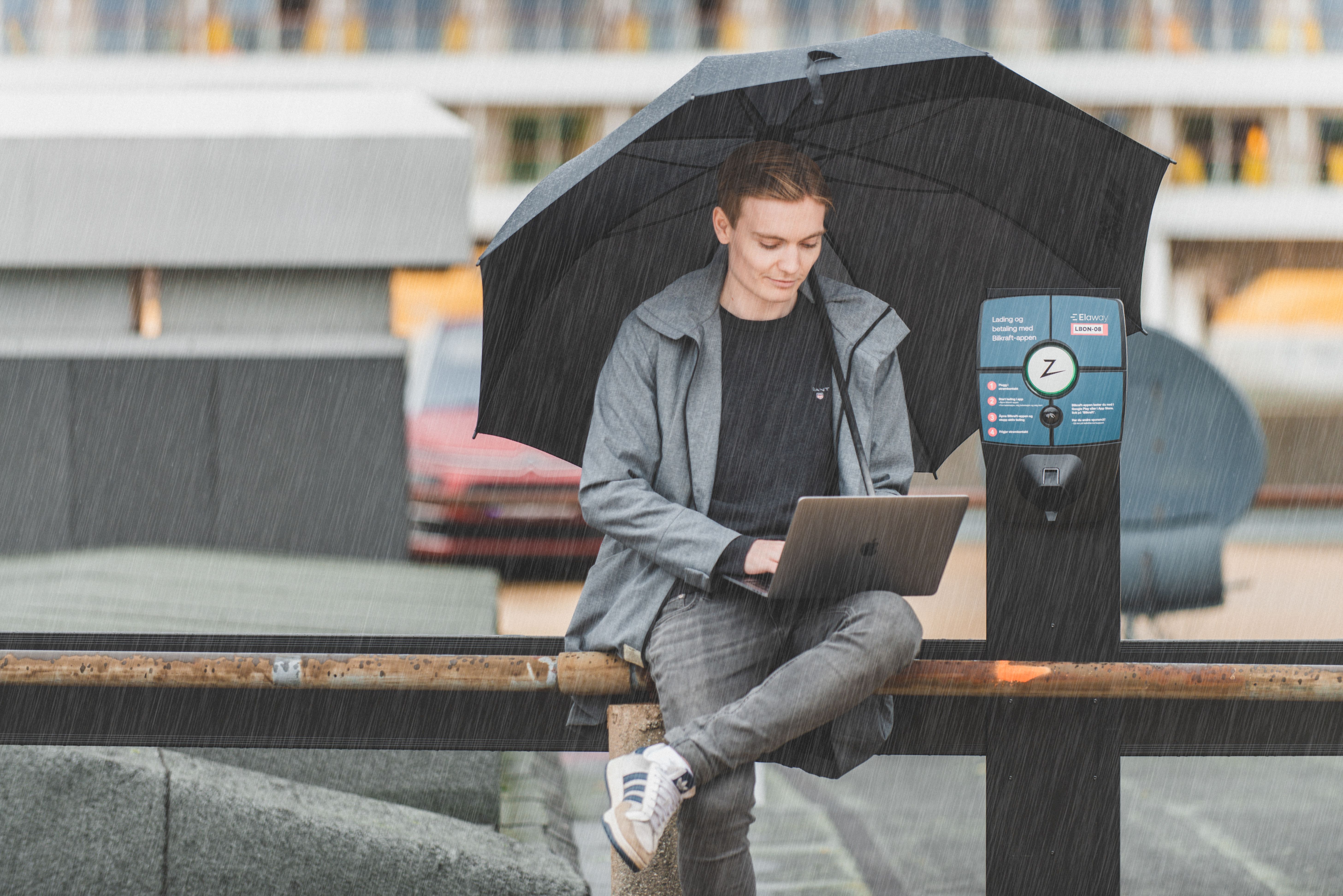 En man sitter på en bänk i regnväder med ett paraply bredvid en Zaptec laddstolpe.