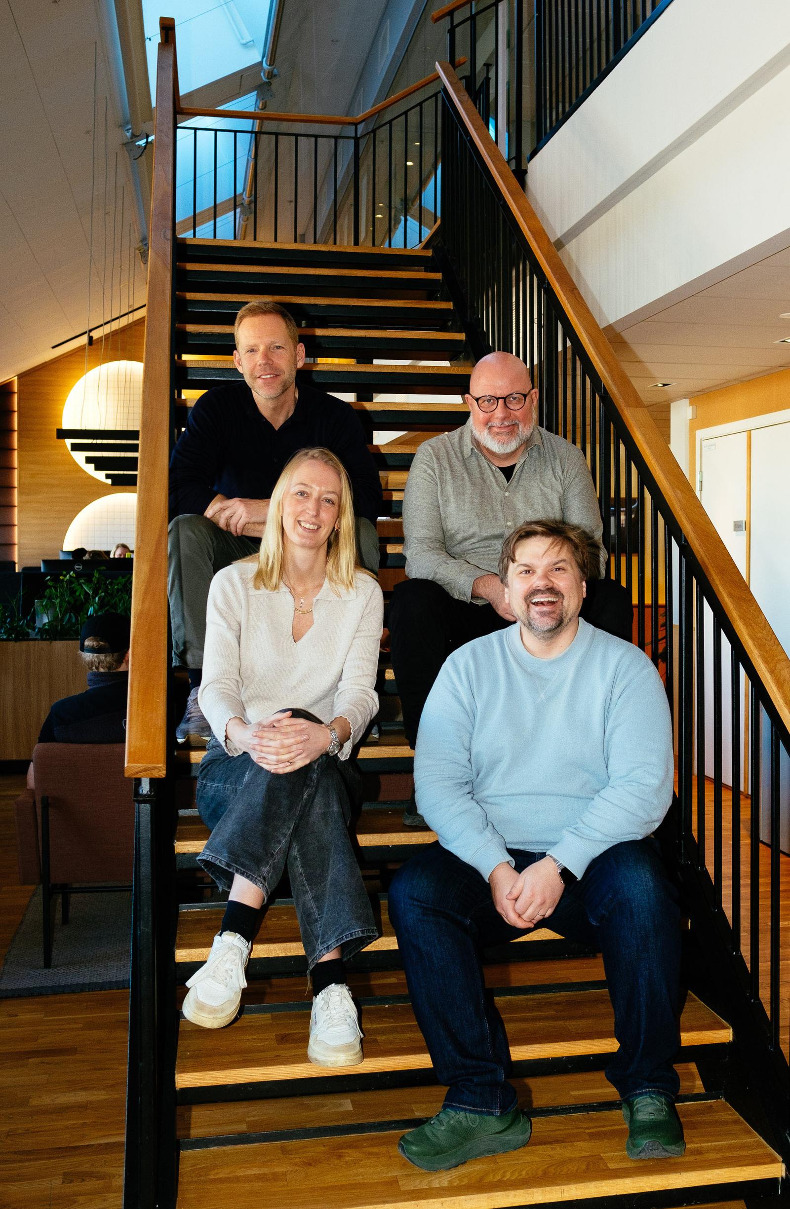 En kvinne og tre menn sitter i en trapp innendørs.
