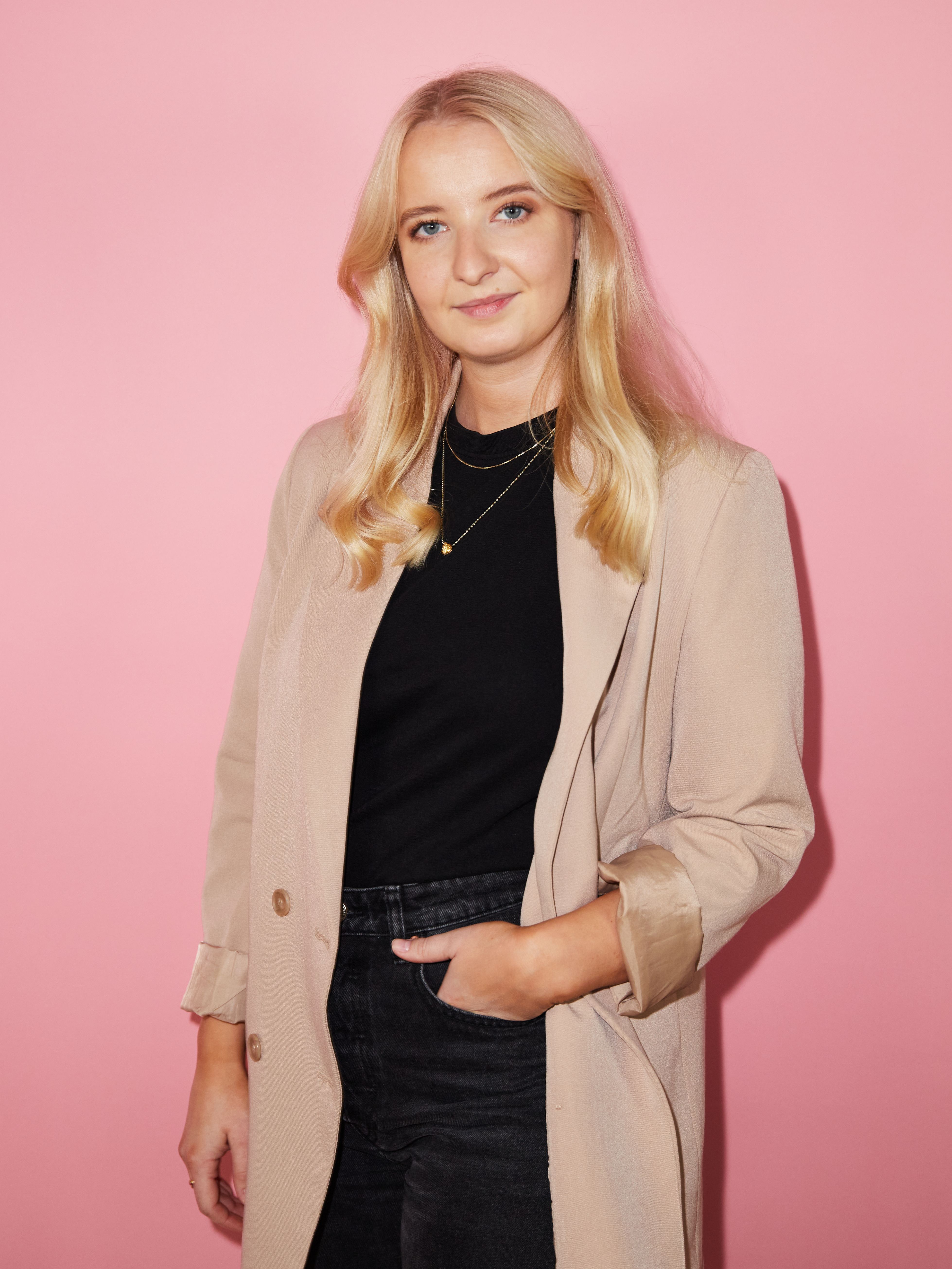 Dame står med hånden i lomma foran en rosa bakgrunn, i beige frakk over sort topp og mørke jeans.