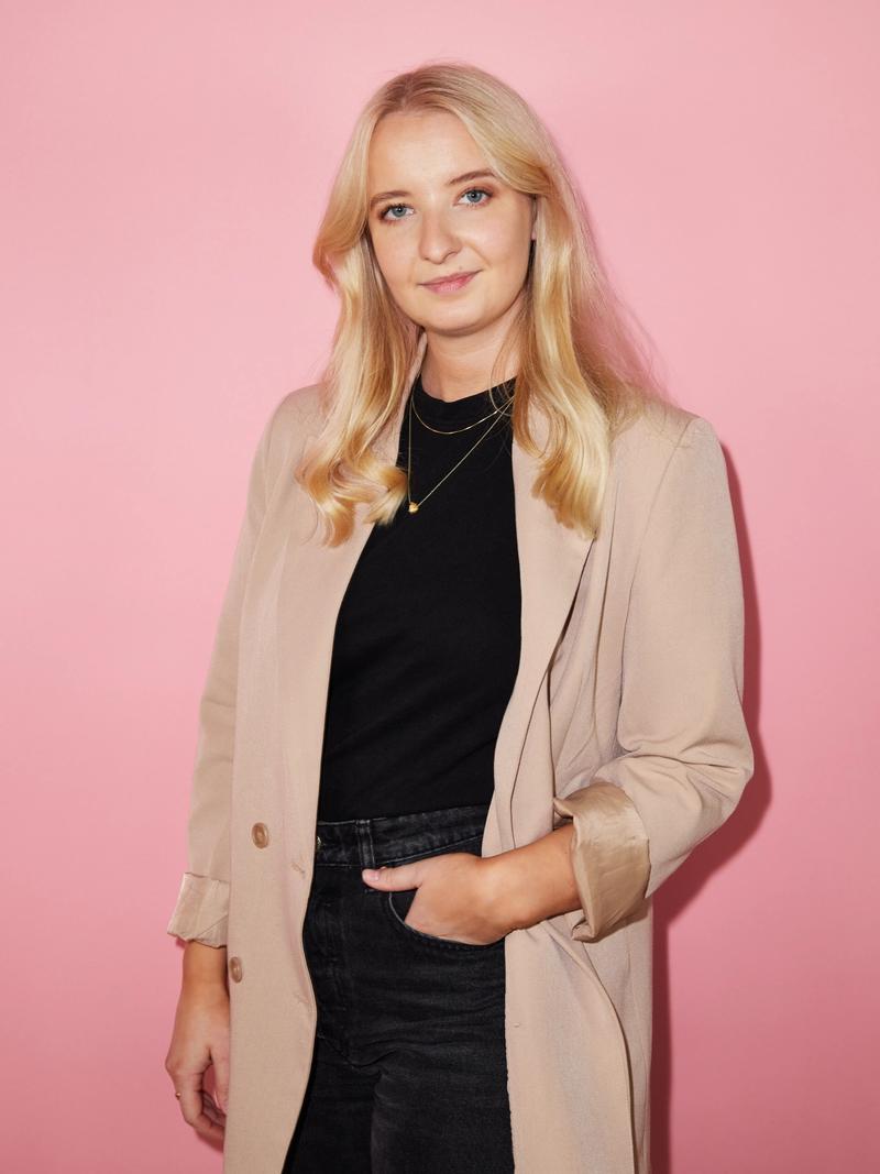 Dame står med hånden i lomma foran en rosa bakgrunn, i beige frakk over sort topp og mørke jeans.