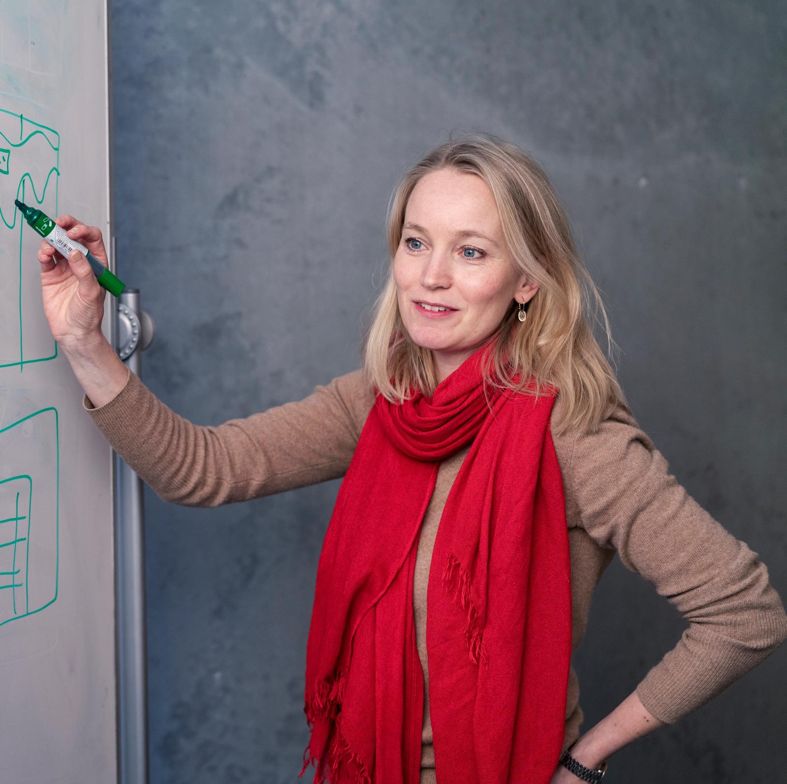Dame med lyst blondt hår og blå øyne skriver på tavle. Hun har på seg et rødt skjerf og beige genser.