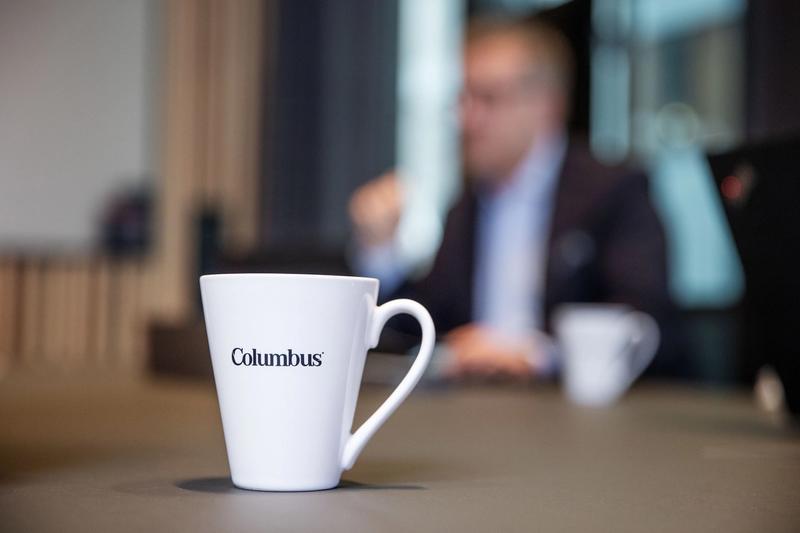 Bilde viser en hvit kaffekopp i fokus, mens i bakgrunnen er det en mann i dress i et møterom på Columbus kontoret