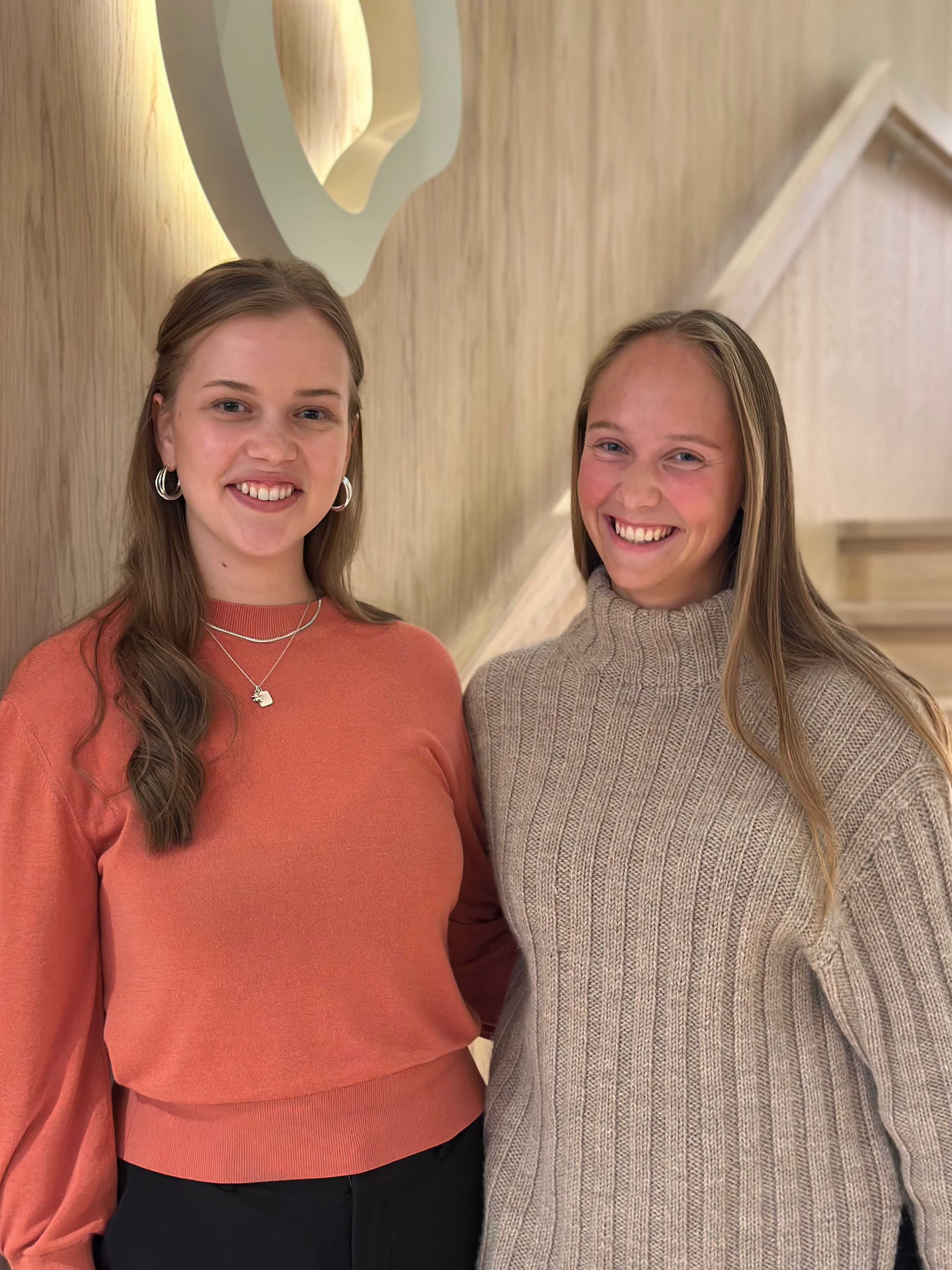 To kvinner står innendørs og smiler.