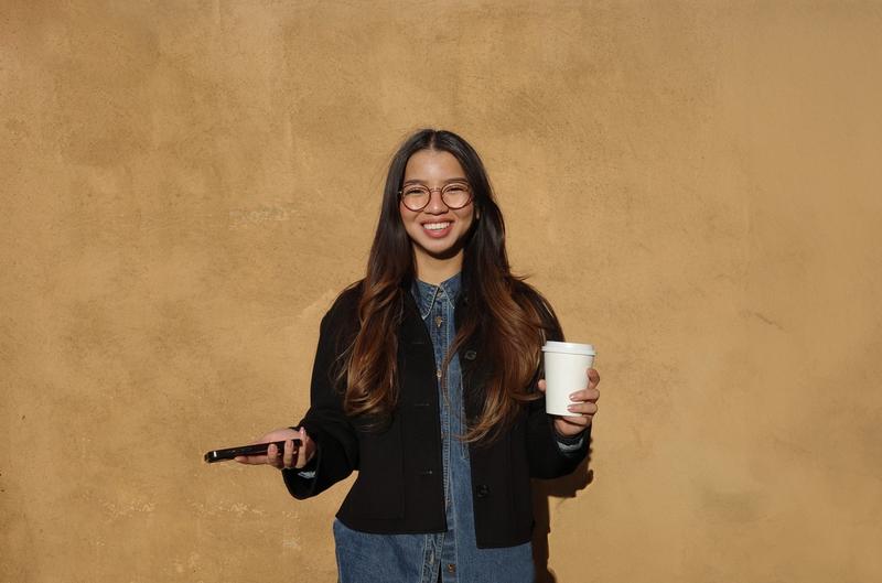 Dame med langt brunt hår og briller. Kaffekopp i den ene hånda og mobilen i den andre, foran en gul vegg utendørs.