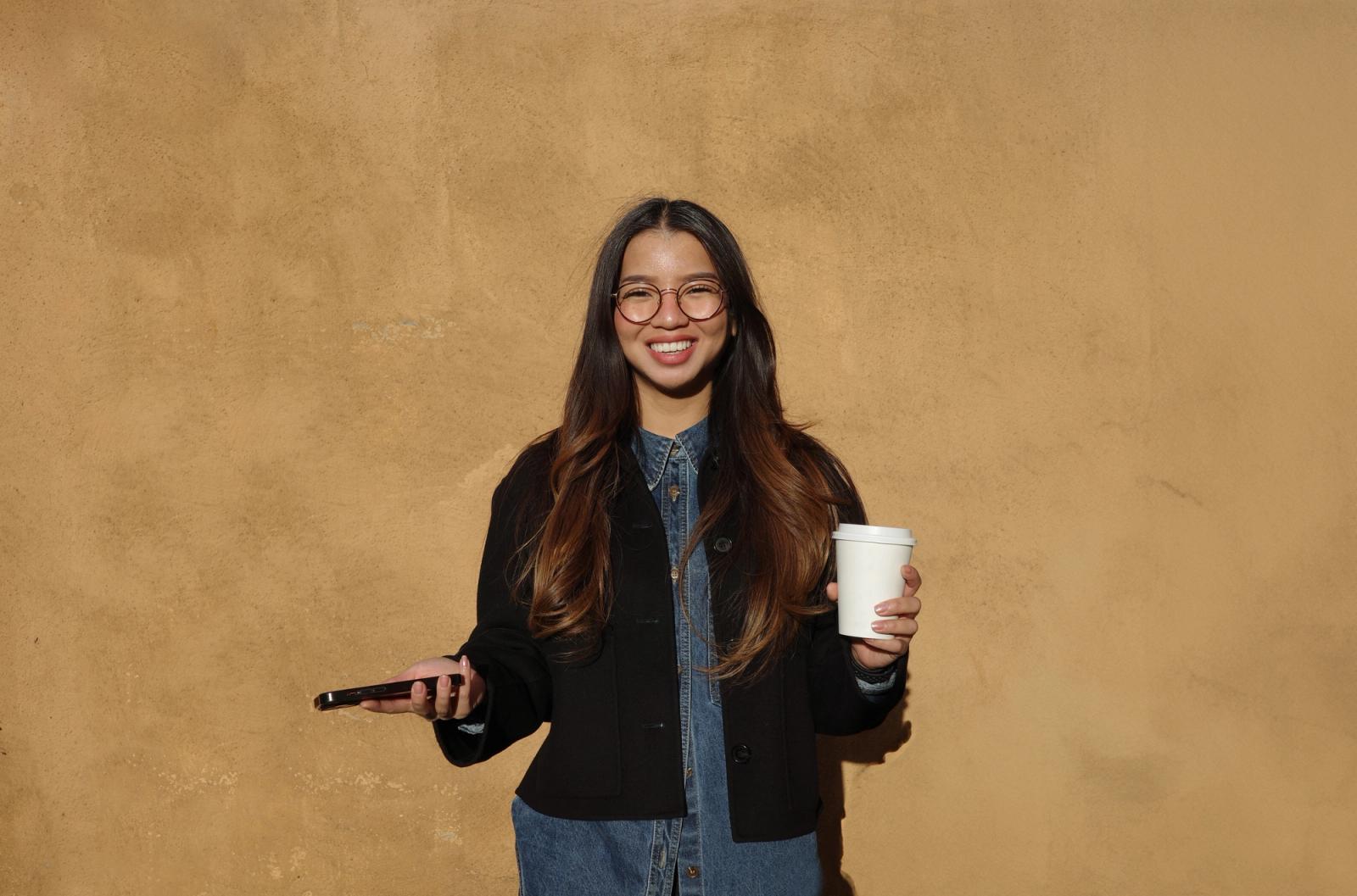 Dame med langt brunt hår og briller. Kaffekopp i den ene hånda og mobilen i den andre, foran en gul vegg utendørs.