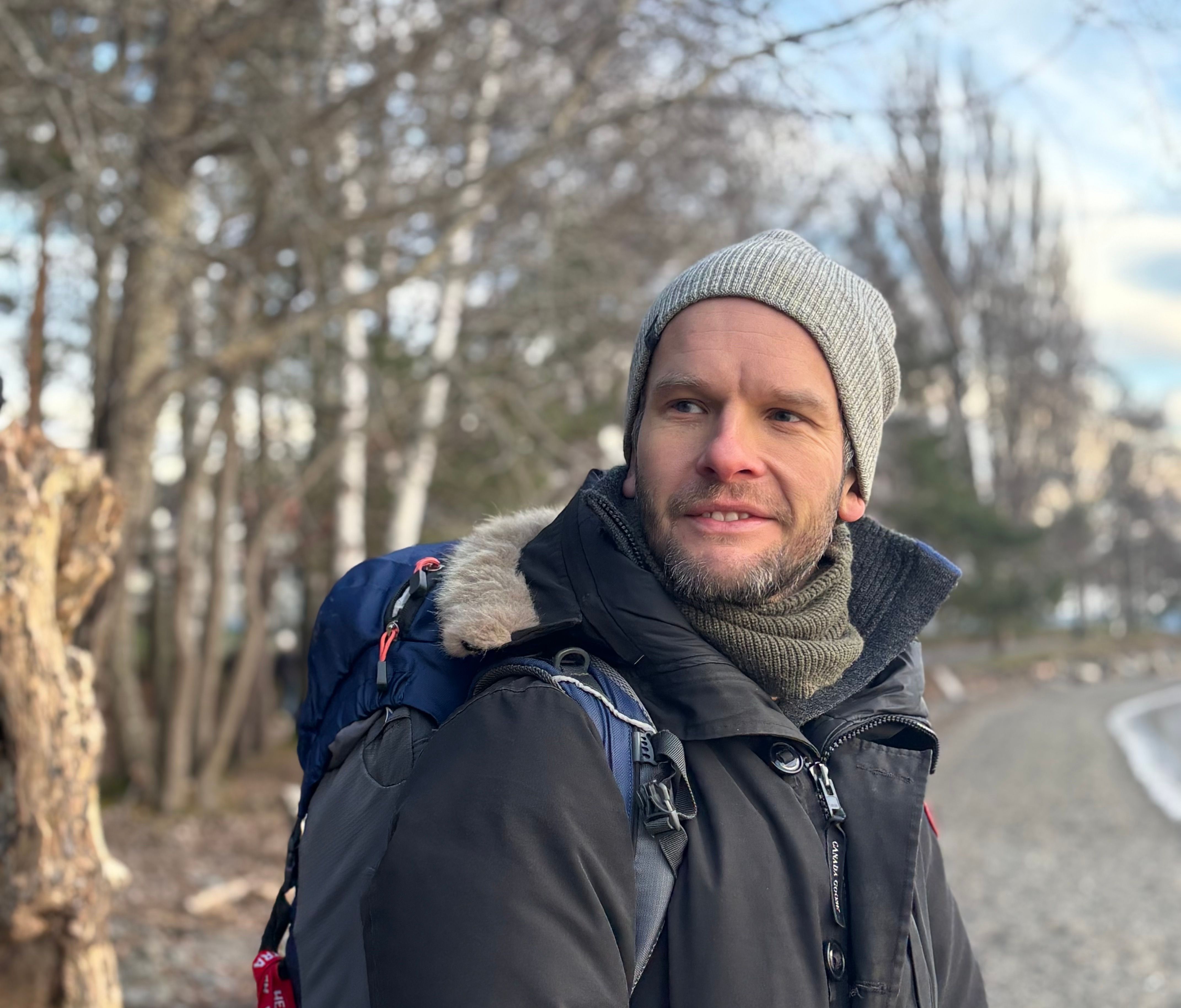 Mann utendørs med sekk, vinterjakke og lue.