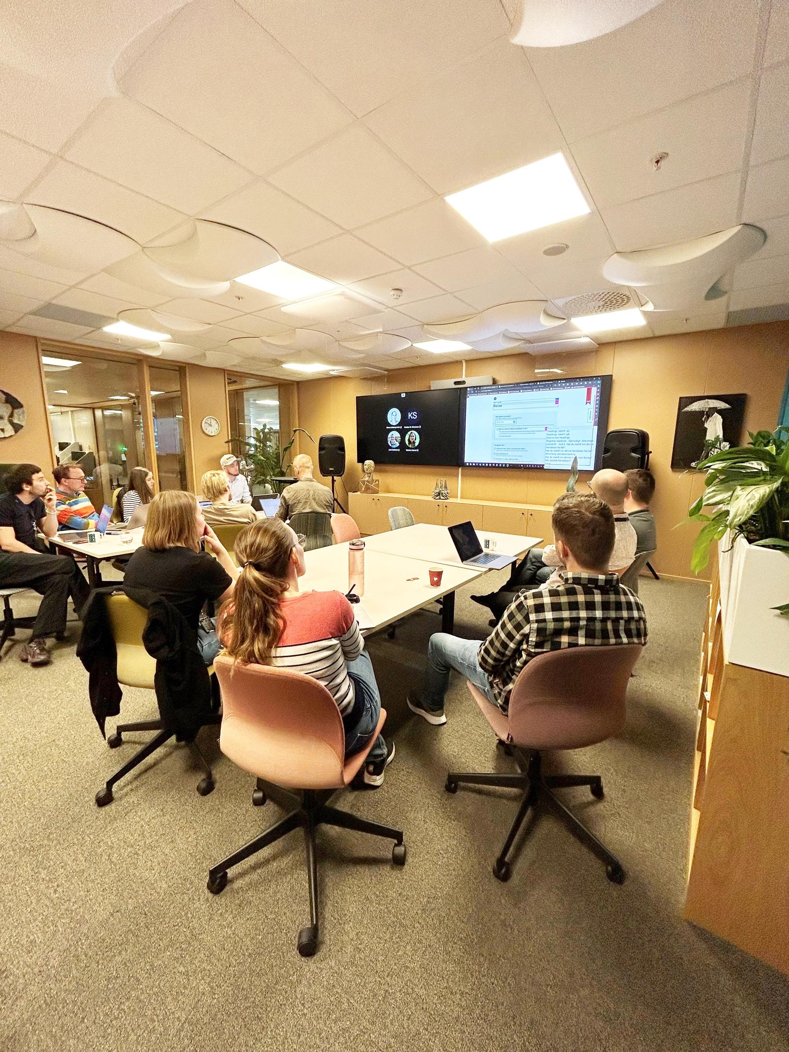 Mennesker sitter rundt bord på et kontor og ser på en skjerm.