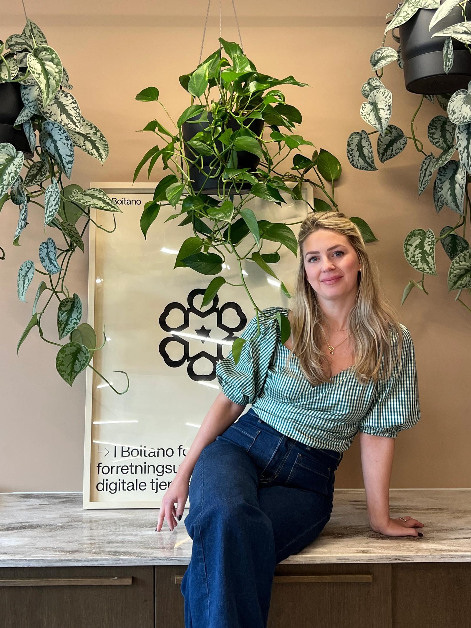 Kvinne med langt lyst hår sitter på en benk, grønne planter henter fra taket.