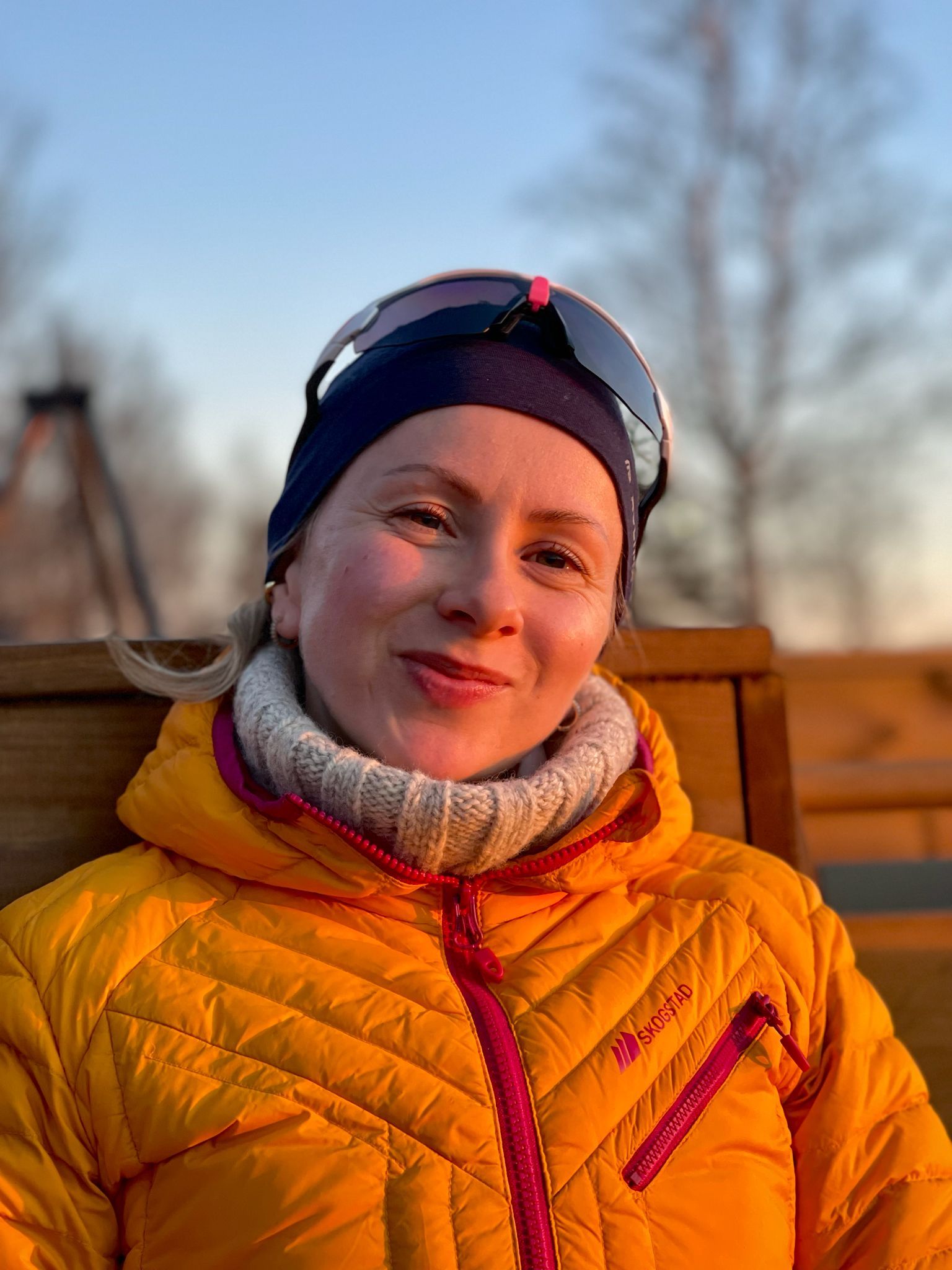 Dame med skibriller, lue og boblejakke sitter utendørs i kveldssola. 