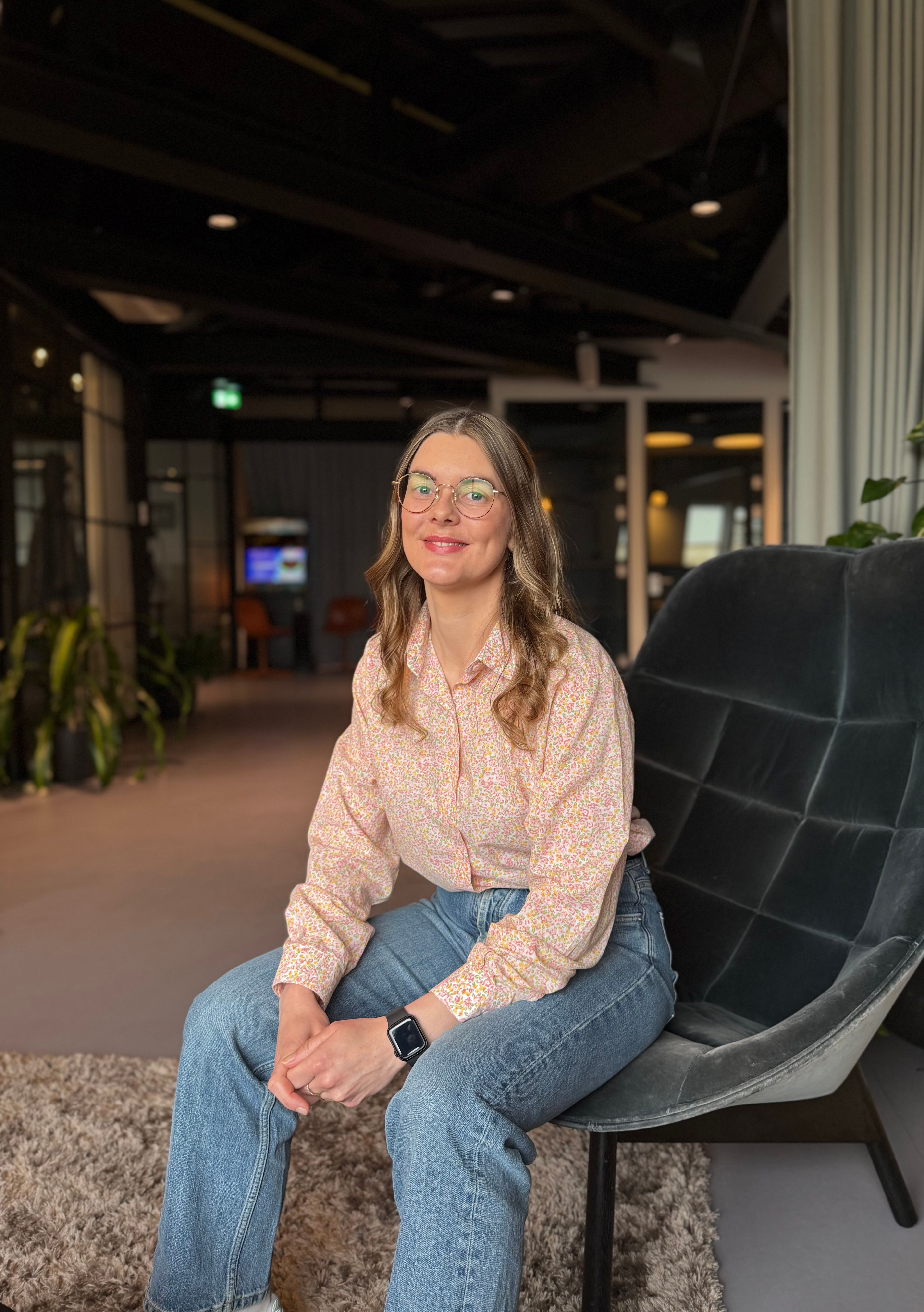 Dame sitter i skinnstol på kontor og smiler. Hun har langt lyst hår og briller. 