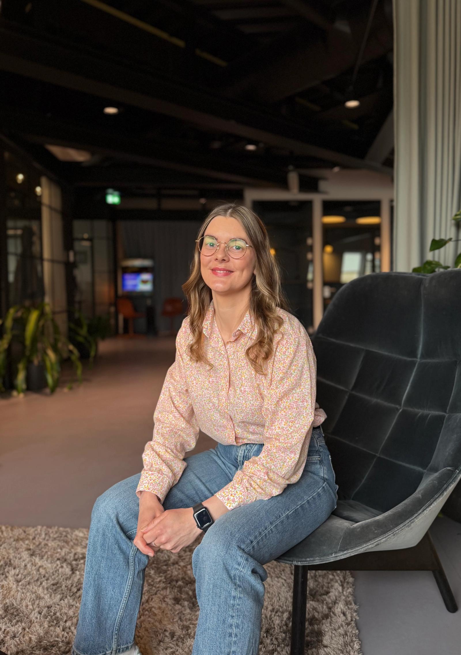 Dame sitter i skinnstol på kontor og smiler. Hun har langt lyst hår og briller.