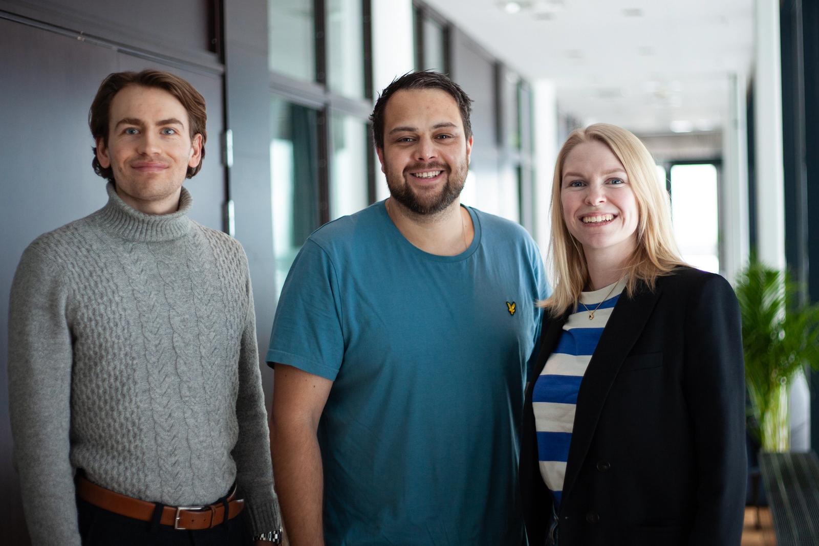 To menn og en kvinne poserer foran kamera i en innendørs gang.