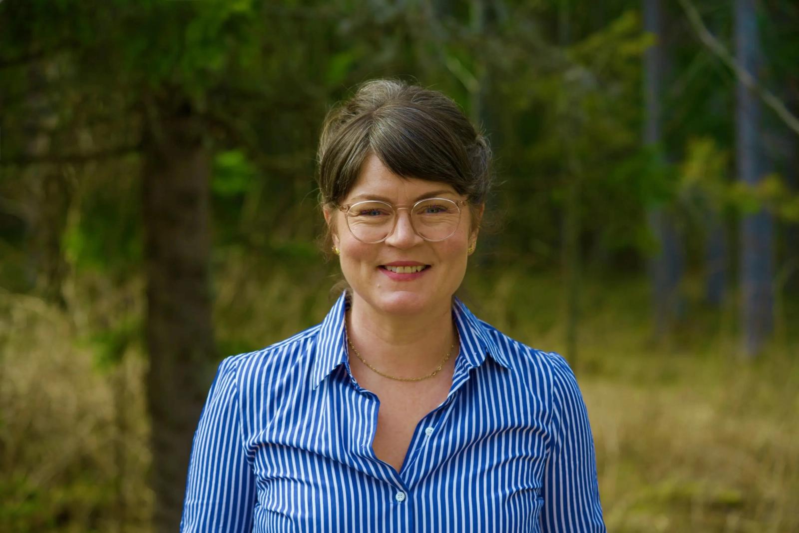 Dame med oppsatt mørkt hår, briller og stripete blå skjorte, ute med skog i bakgrunnen.