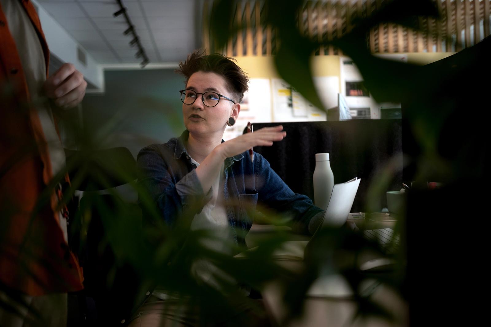 Dame med kort hår, briller og blå skjorte sitter og snakker med person som står ved siden av henne. Bildet er tatt gjennom en plante som har stort fokus i forgrunnen.