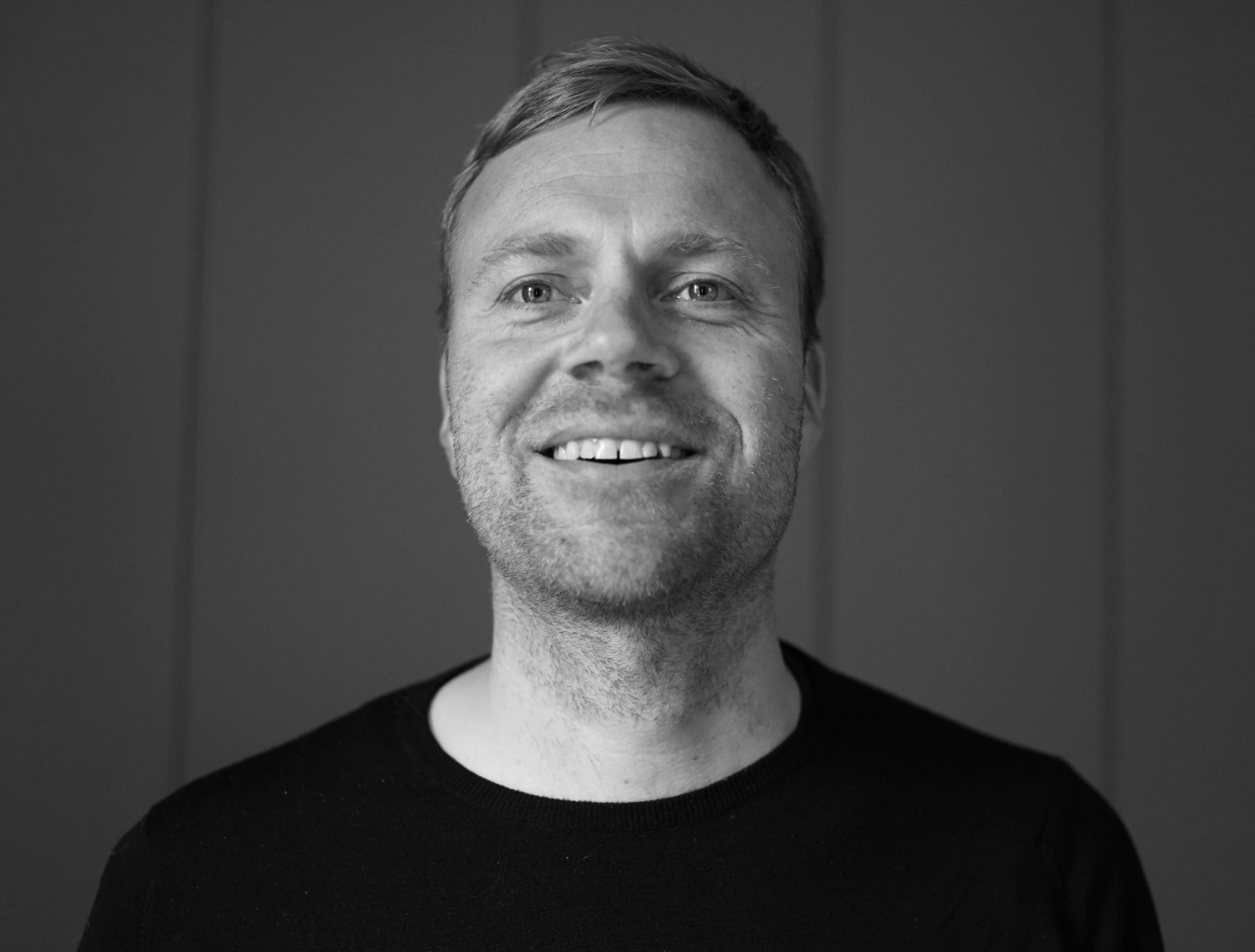 Mann med svart genser poserer mot grå bakgrunn. Bildet er svart-hvit.