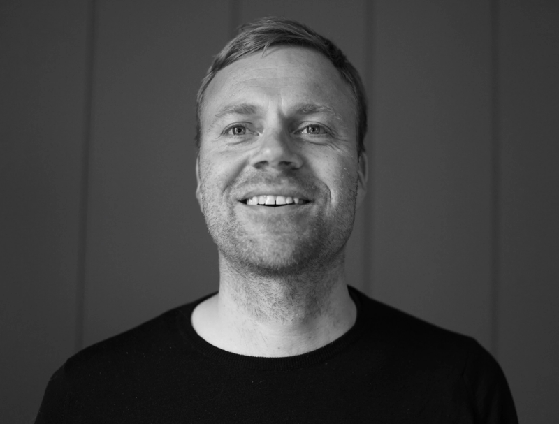 Mann med svart genser poserer mot grå bakgrunn. Bildet er svart-hvit.