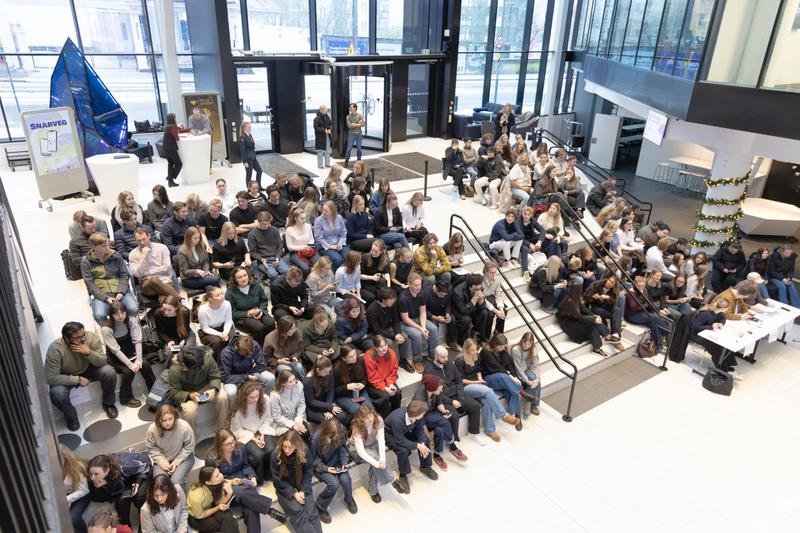 En stor gjeng mennesker sitter i trapper eller benker i en stor sal