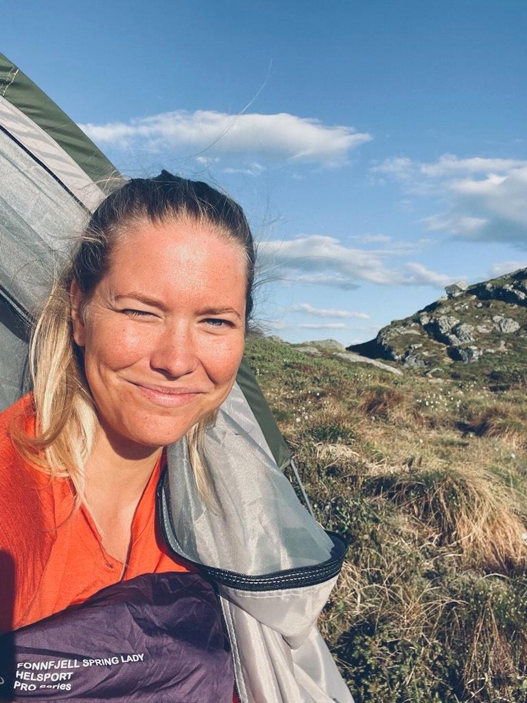 Dame med lyst blondt hår stikker hodet ut i fra telt. Hun smiler og myser med øynene mot solskinnet.
