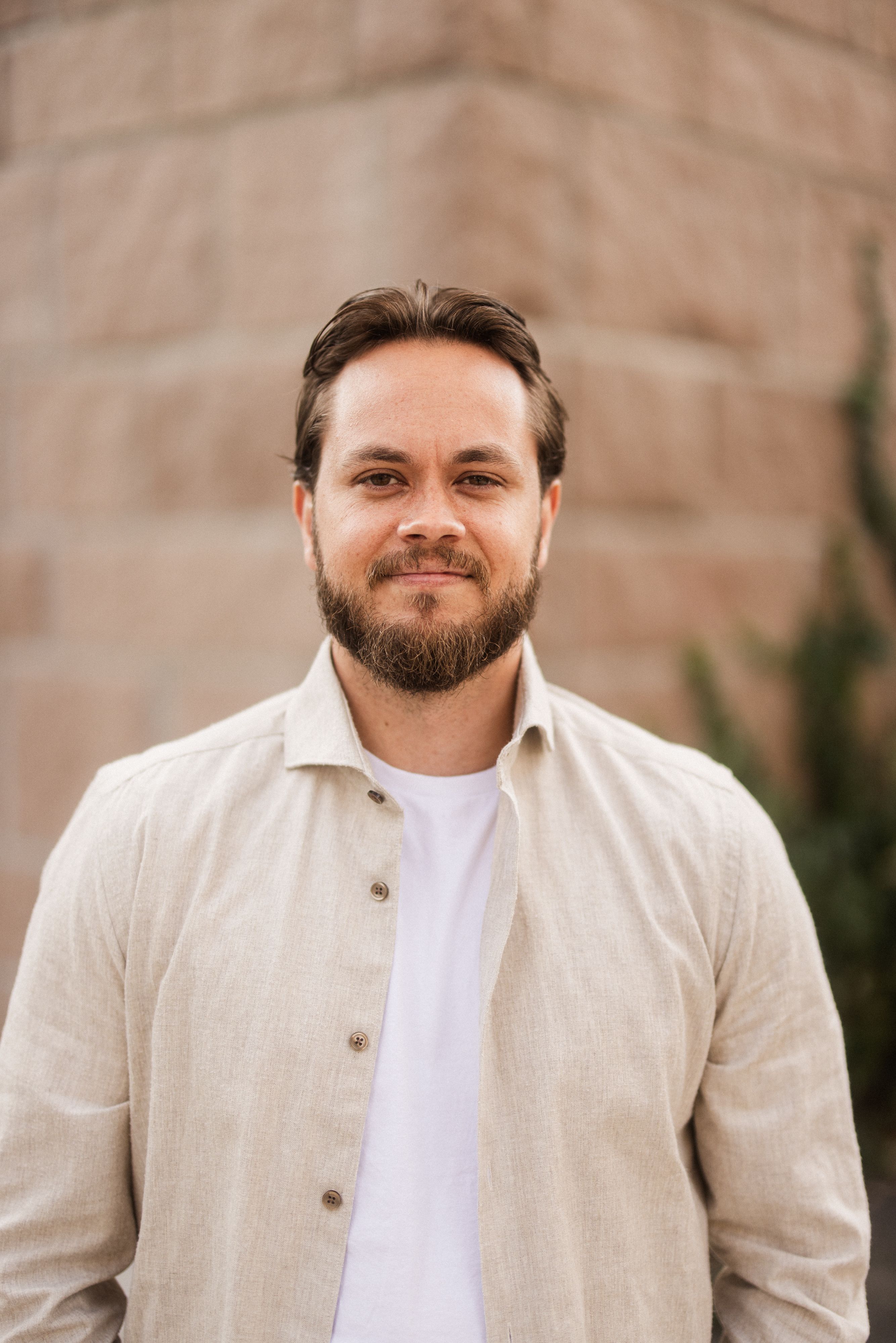 Mann med mørkt hår og kort skjegg, hvit t-skjorte med åpen beige skjorte over