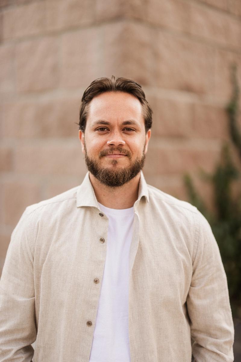 Mann med mørkt hår og kort skjegg, hvit t-skjorte med åpen beige skjorte over