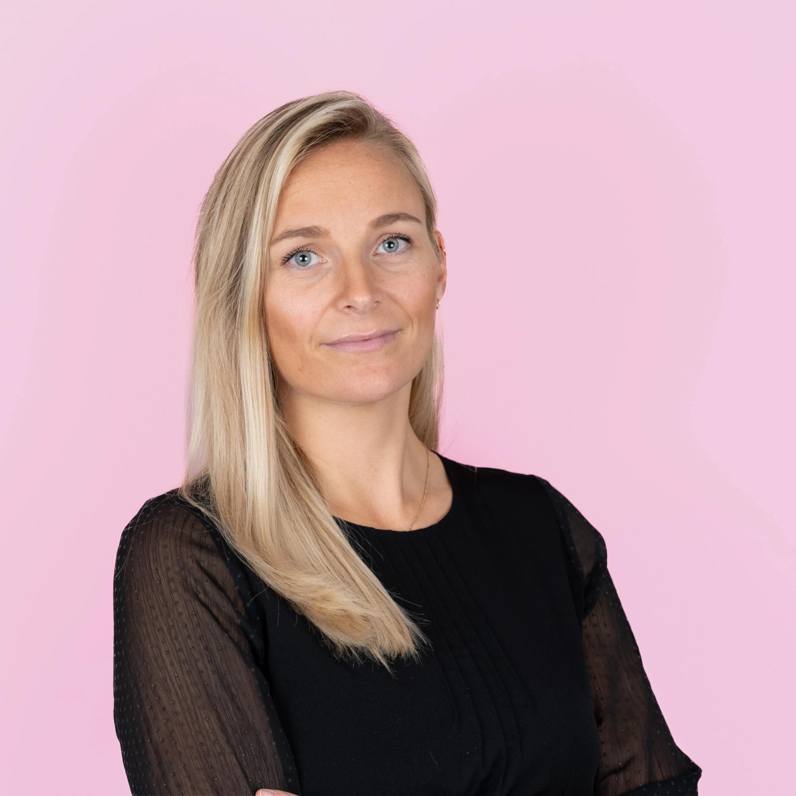 Blond dame med svart skjorte mot rosa bakgrunn.