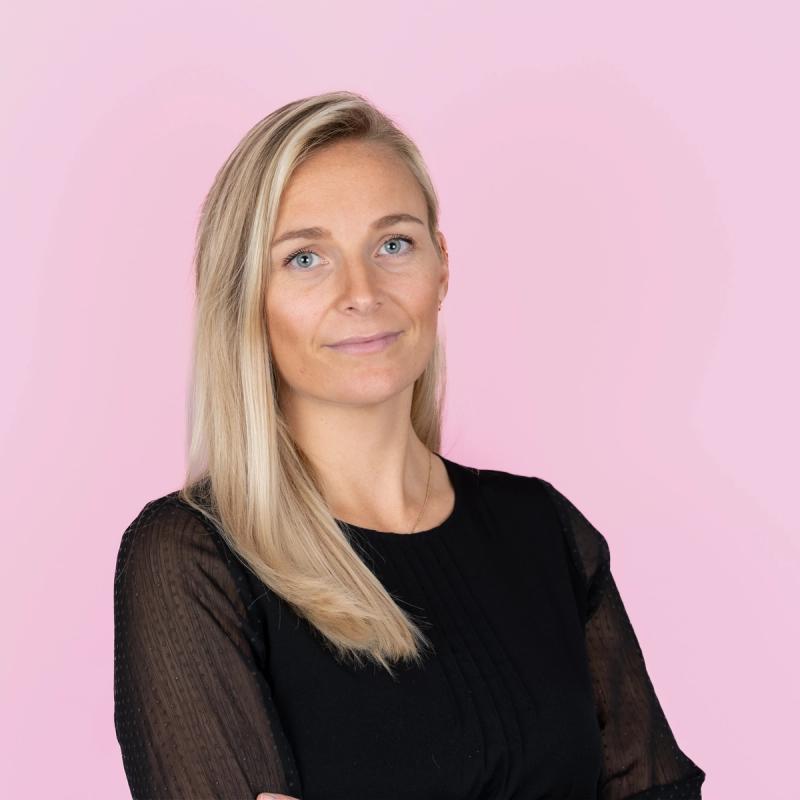 Blond dame med svart skjorte mot rosa bakgrunn.