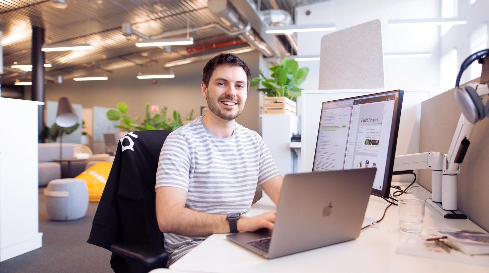 Man sitter på skrivepult i kontorlanskap med laptop og tilkoblet PC-skjerm og smiler til kamera i stripete t-skjorte.
