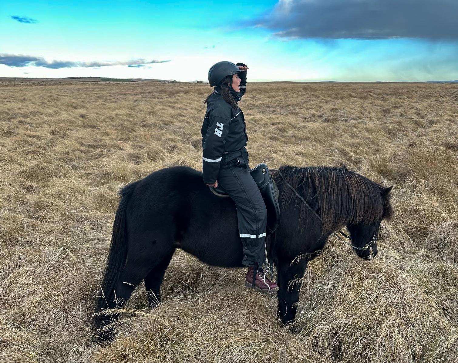 Dame i kjeledress og hjem sitter på en svart islandshest ute på et stort jorde.