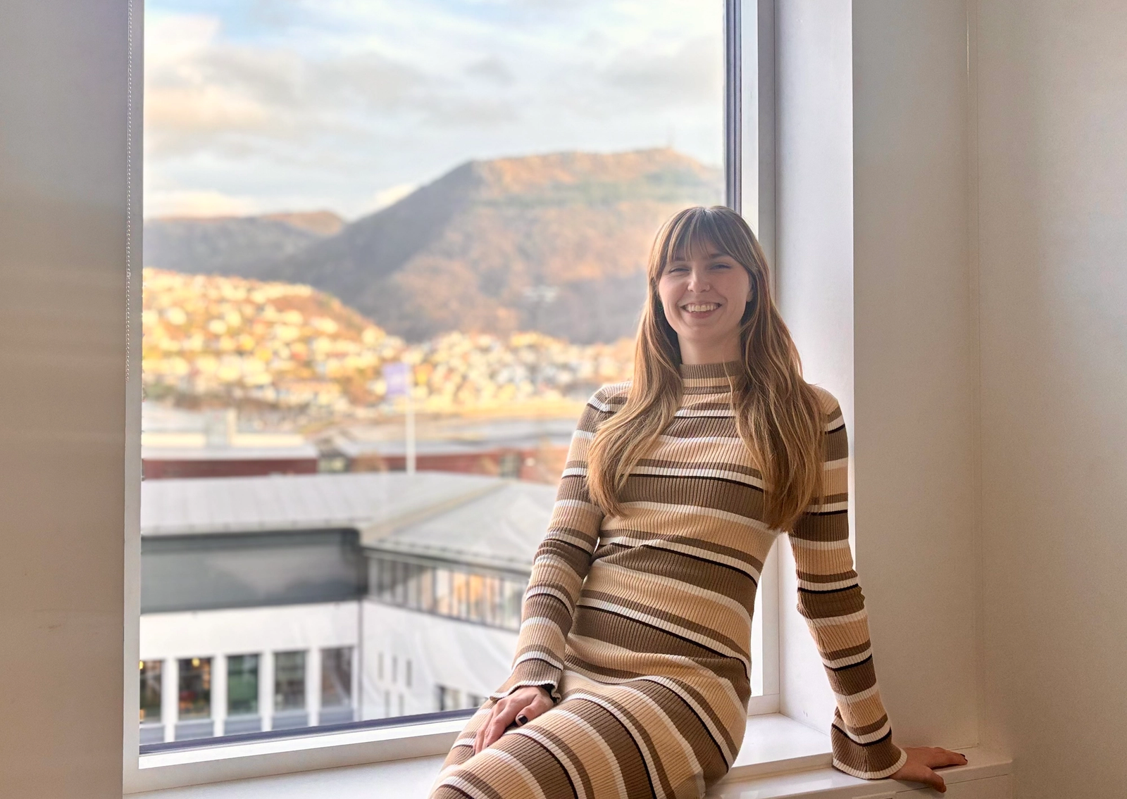 Dame i stripete kjole sitter i vinduskarm med Bergen og Ulrikken i bakgrunnen.