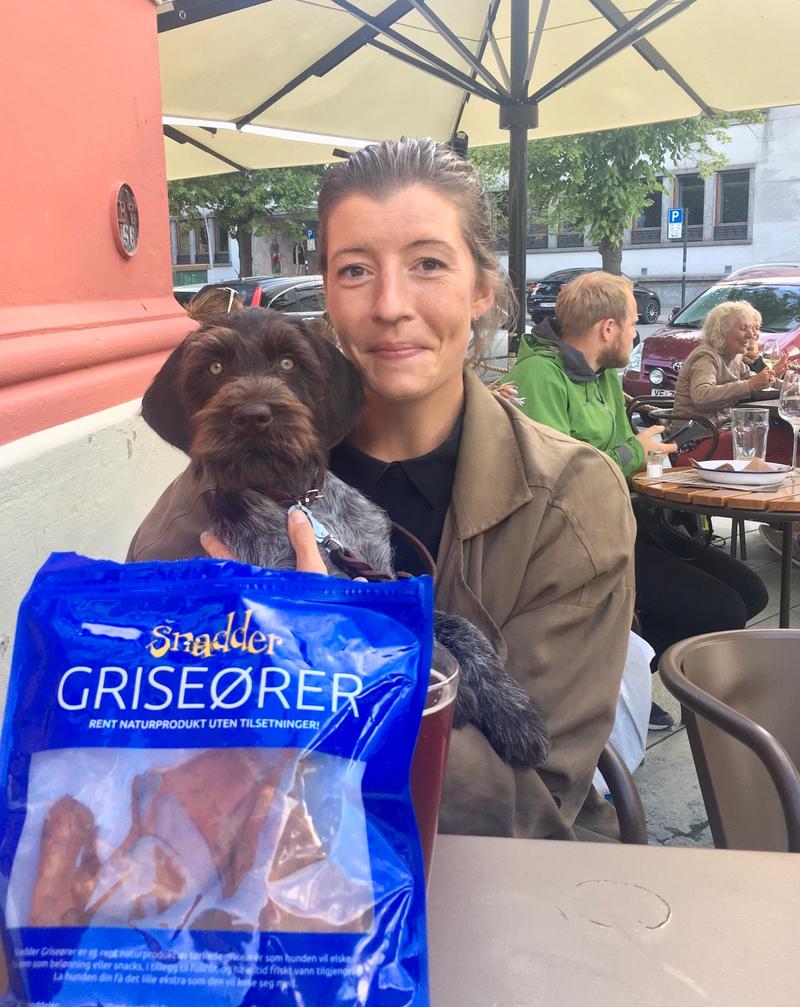 Dame sitter på café med hund på fanget. Framfor dem er det en pose med griseører.
