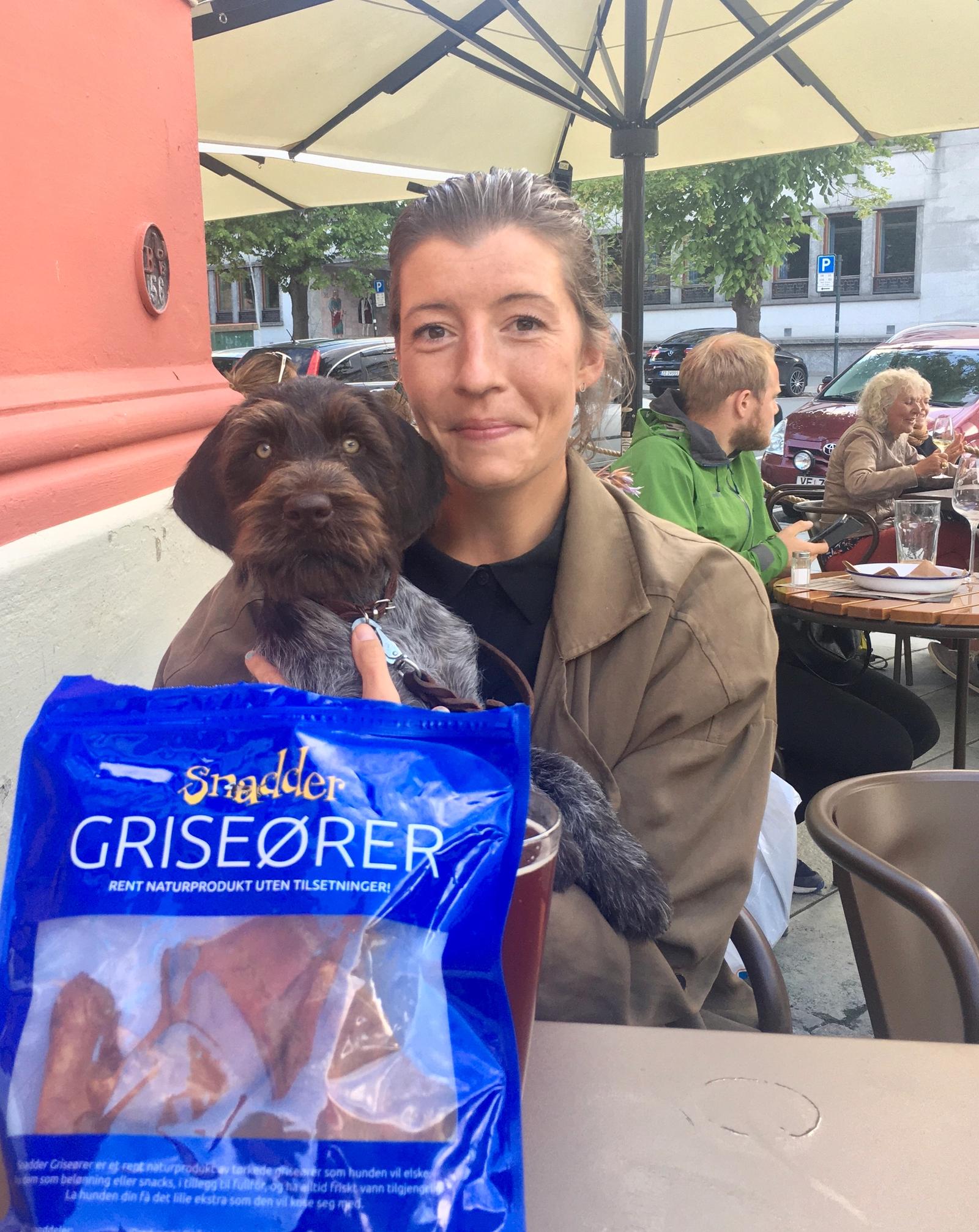 Dame sitter på café med hund på fanget. Framfor dem er det en pose med griseører.
