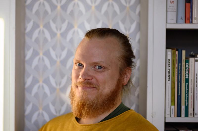 Mann med blå øyne, langt hår i knute og skjegg. I bakgrunnen er det hyller med bøker.