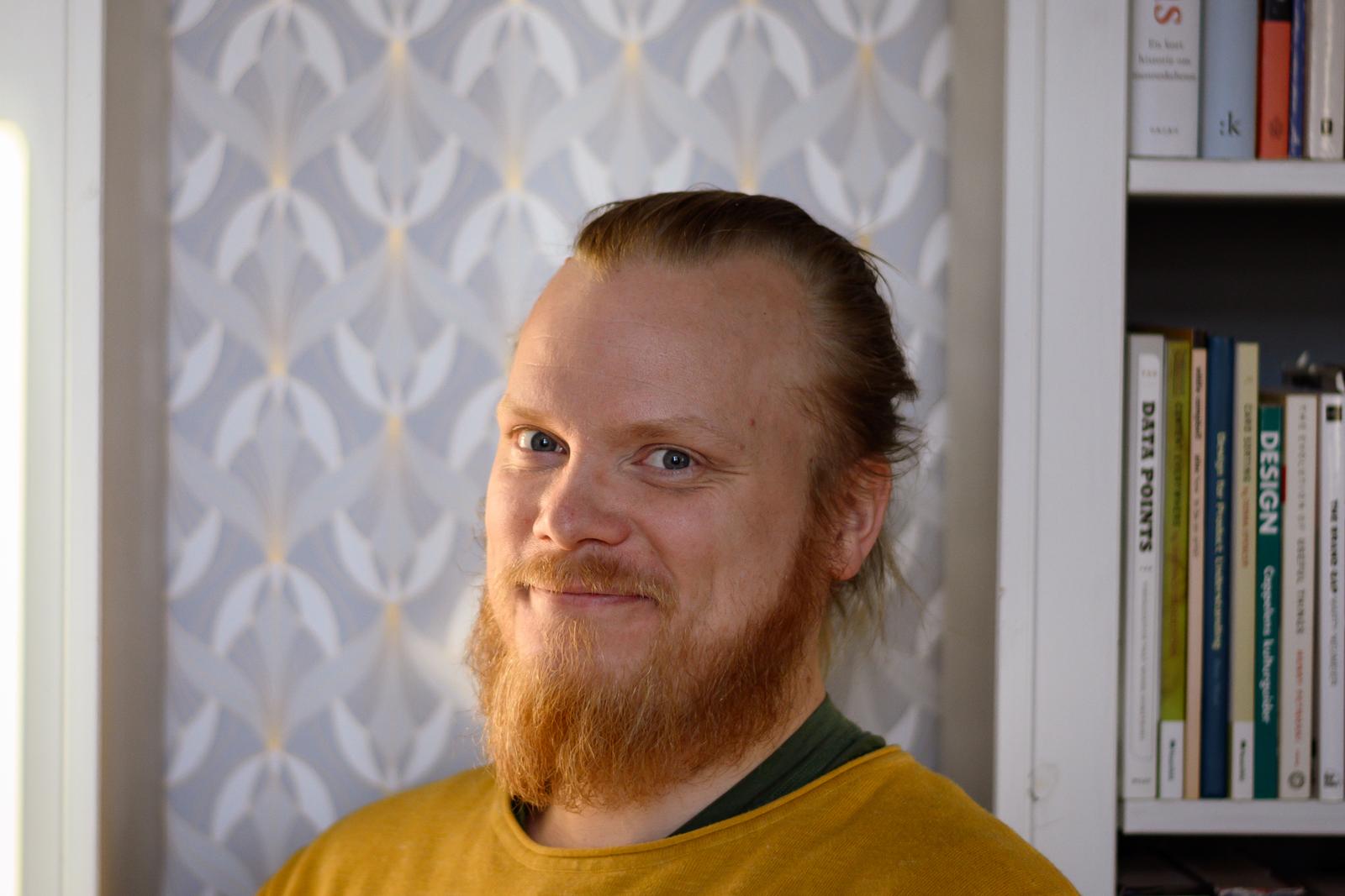 Mann med blå øyne, langt hår i knute og skjegg. I bakgrunnen er det hyller med bøker.