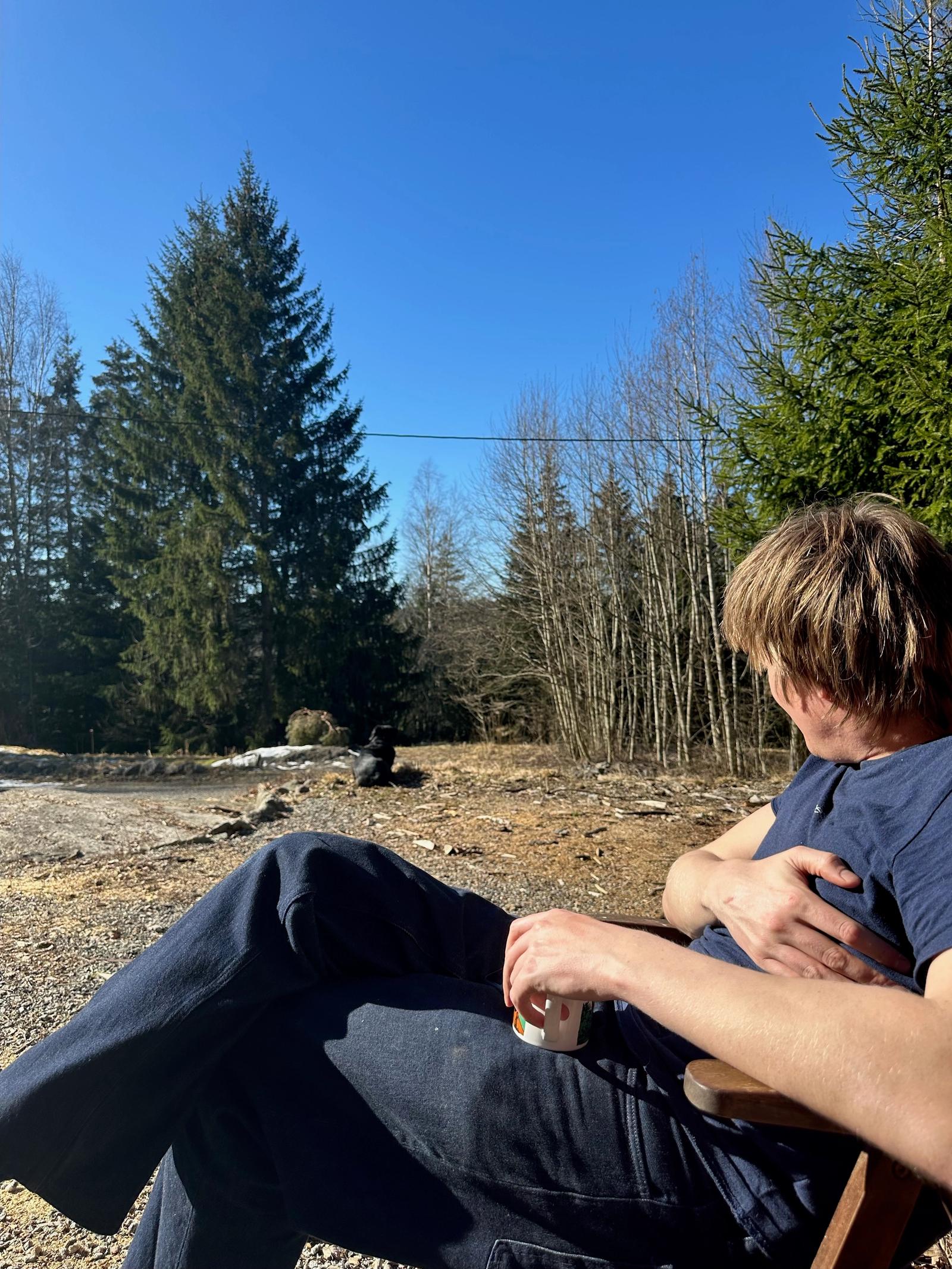 Mann sitter i stol utendørs med drikke på boks og ser mot en hund.