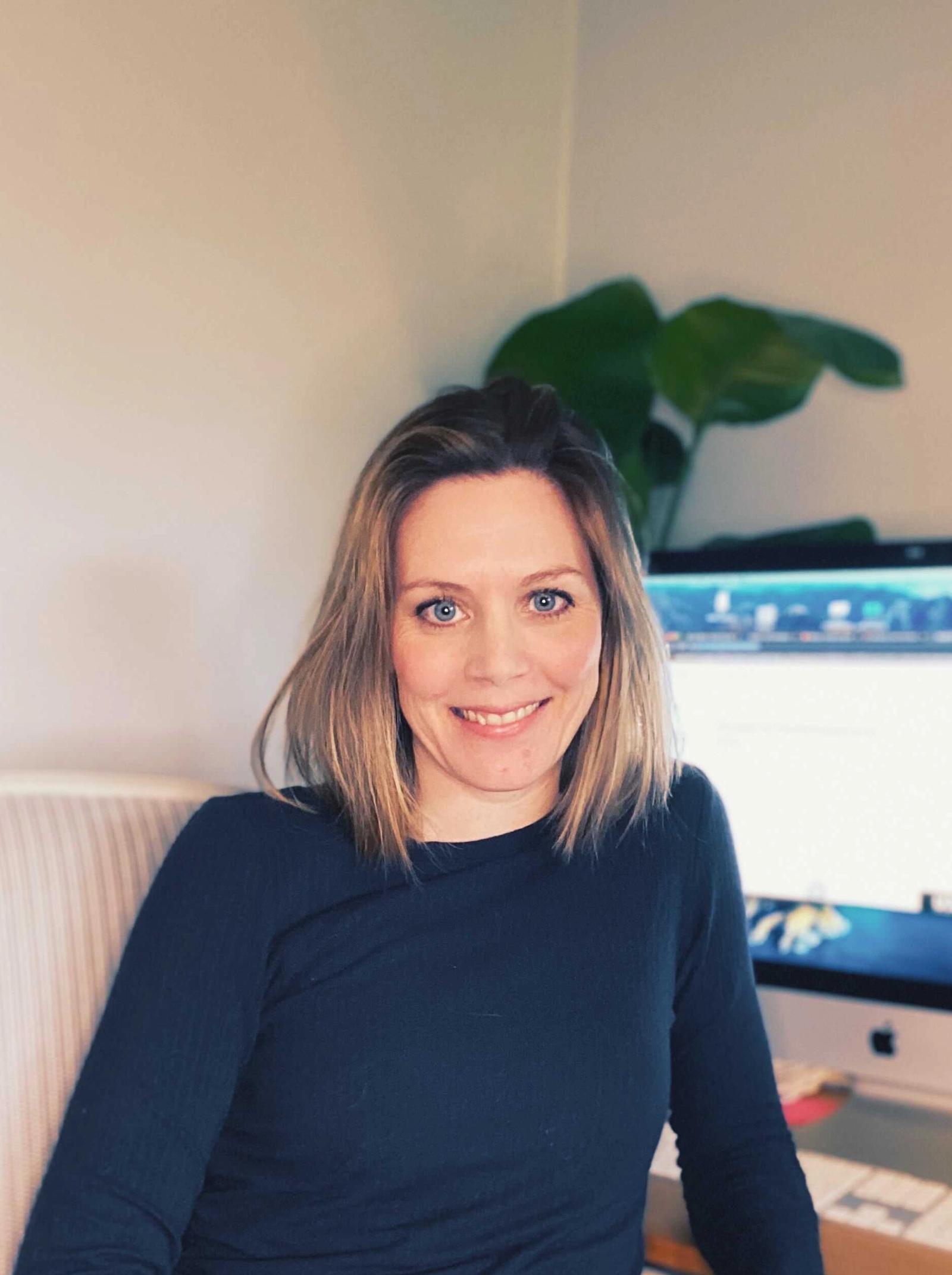 Dame med blå øyne og brunt hår med blonde highlights til skuldrene sitter med PC. Hun har på seg en enkel svart top.