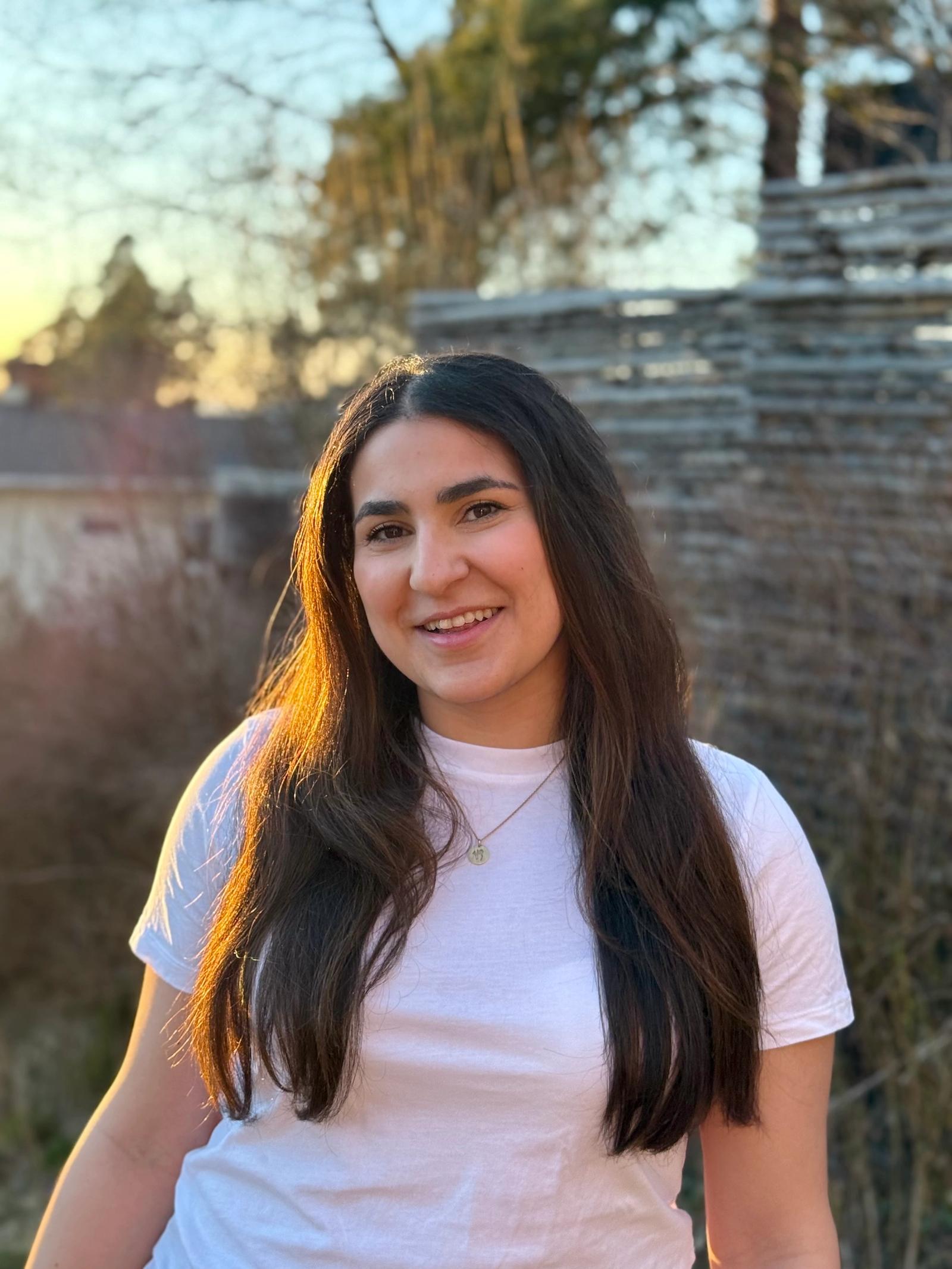 Dame med mørkt langt hår og hvit t-skjorte står utendørs i kveldssol og smiler.