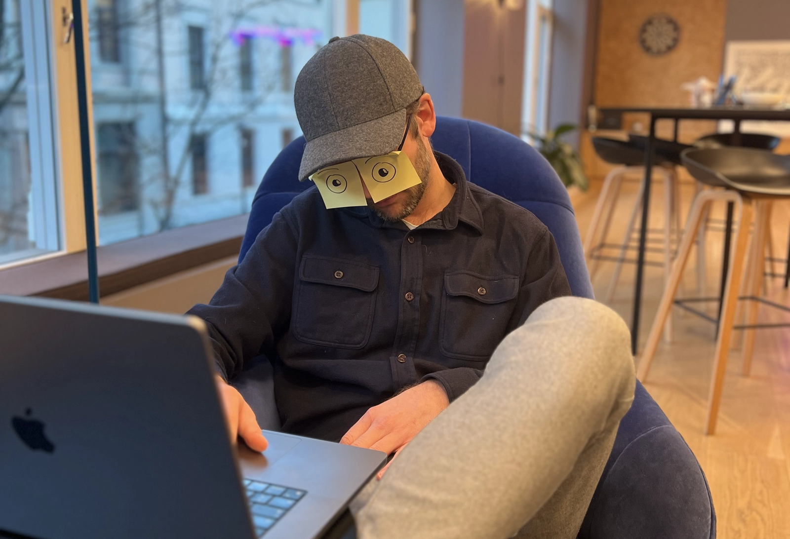 Mann sitter i en stol med laptopen foran seg. Har post-it-lapper på øynene og ser ut som om han sover.