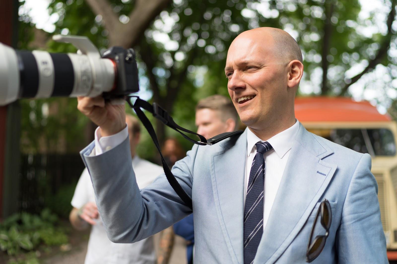 Mann i blÄ dress med grÄtt, stripete slips stÄr med kamera i hÄnden.