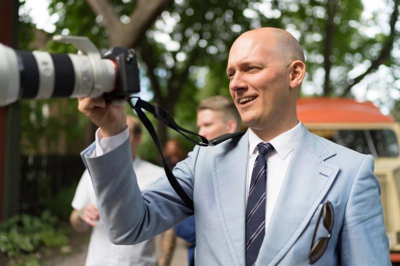 Mann i blå dress med grått, stripete slips står med kamera i hånden.