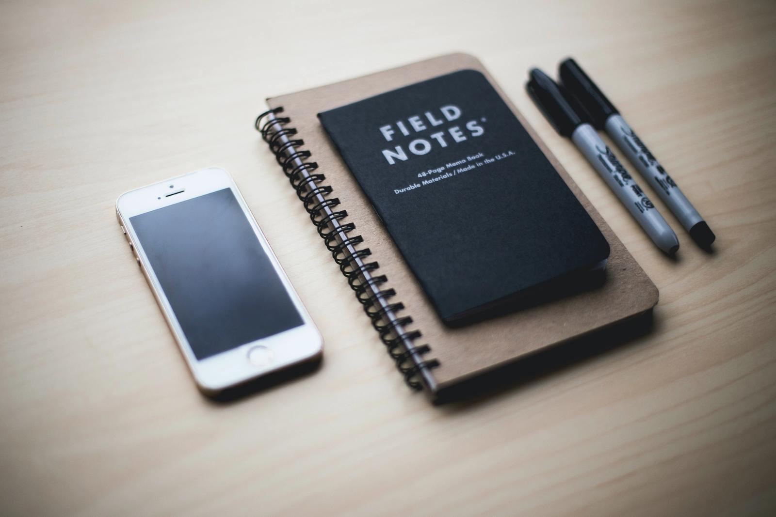 En mobil, notatbok og to penner ligger på et bord.