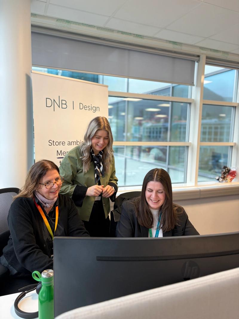 To kvinner sitter ved et skrivebord med en skjerm på, en tredje kvinne står bak dem.