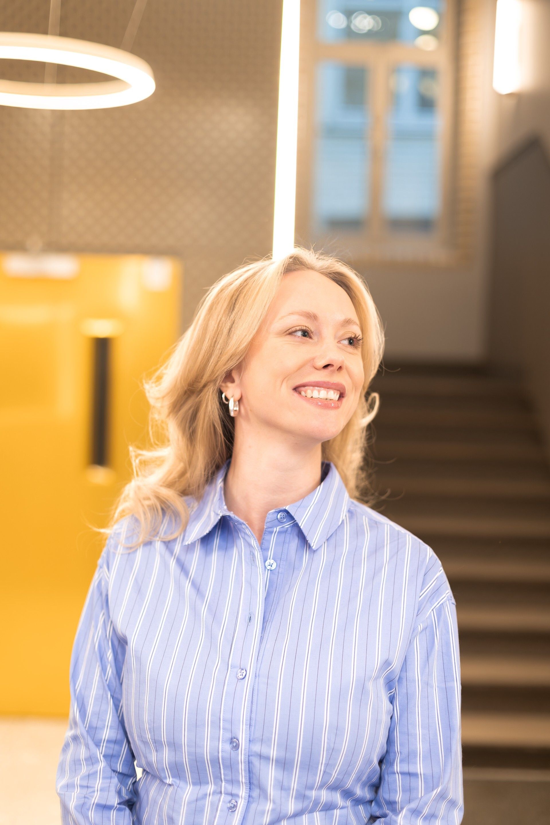 Dame med blondt hår og dongeri-skjorte poserer med bildevegg sammen med to andre personer. 